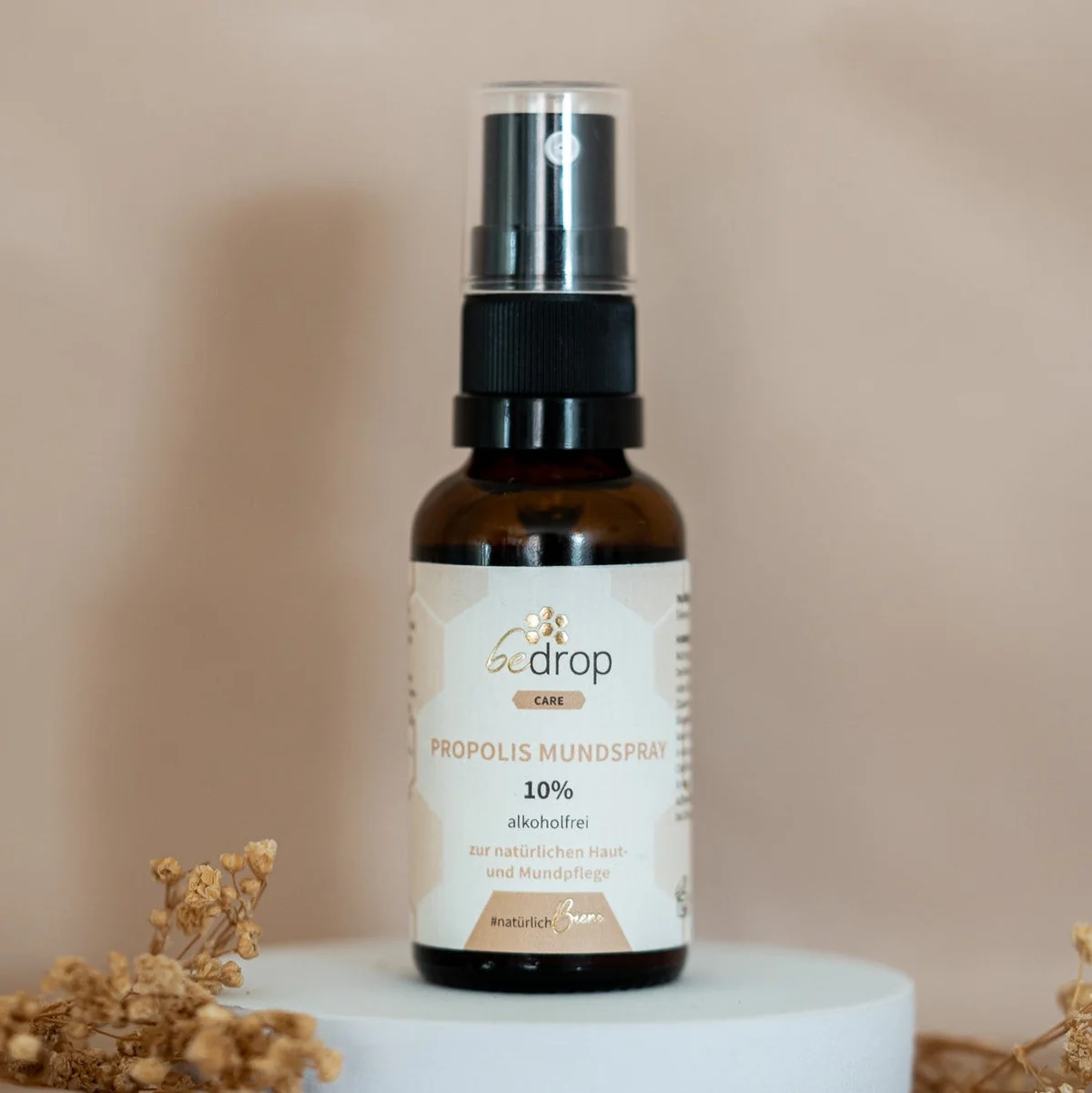 bedrop Propolis Mundspray 10 % alkoholfrei in brauner Glasflasche mit schwarzem Sprühkopf und weiß-beigem Etikett, dekorativ auf weißem Podest mit Trockenblumen präsentiert.