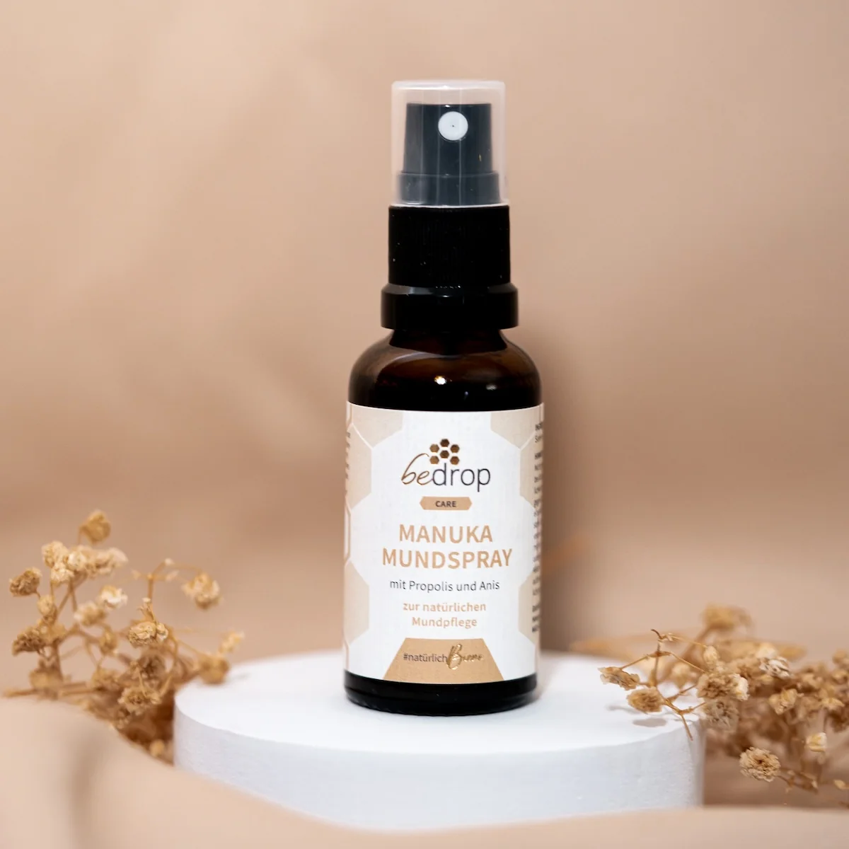 bedrop Manuka Mundspray mit Propolis und Anis in brauner Glasflasche mit schwarzem Sprühkopf, dekorativ präsentiert auf weißem Podest mit Trockenblumen